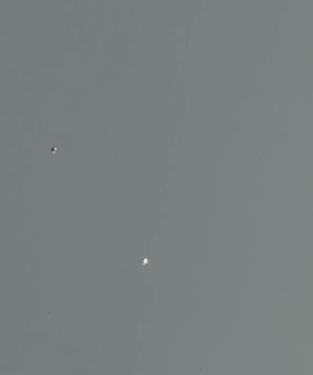 Enlarged detail of the two objects from the same frame. Neither resolves into a recognisable aircraft, balloon, or bird at this distance against the overcast.
