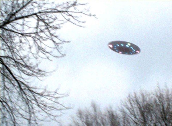 Green Bay, Wisconsin, first week of January 2007: one of two photos of a dark disc with a ring of multicolored underside lights, shot through bare winter trees. Submitted to UFOEvidence.org by investigator Jack Nethering on behalf of an anonymous couple, and widely treated as a companion to the 2003 Weyauwega frames because the craft looks like the same object.